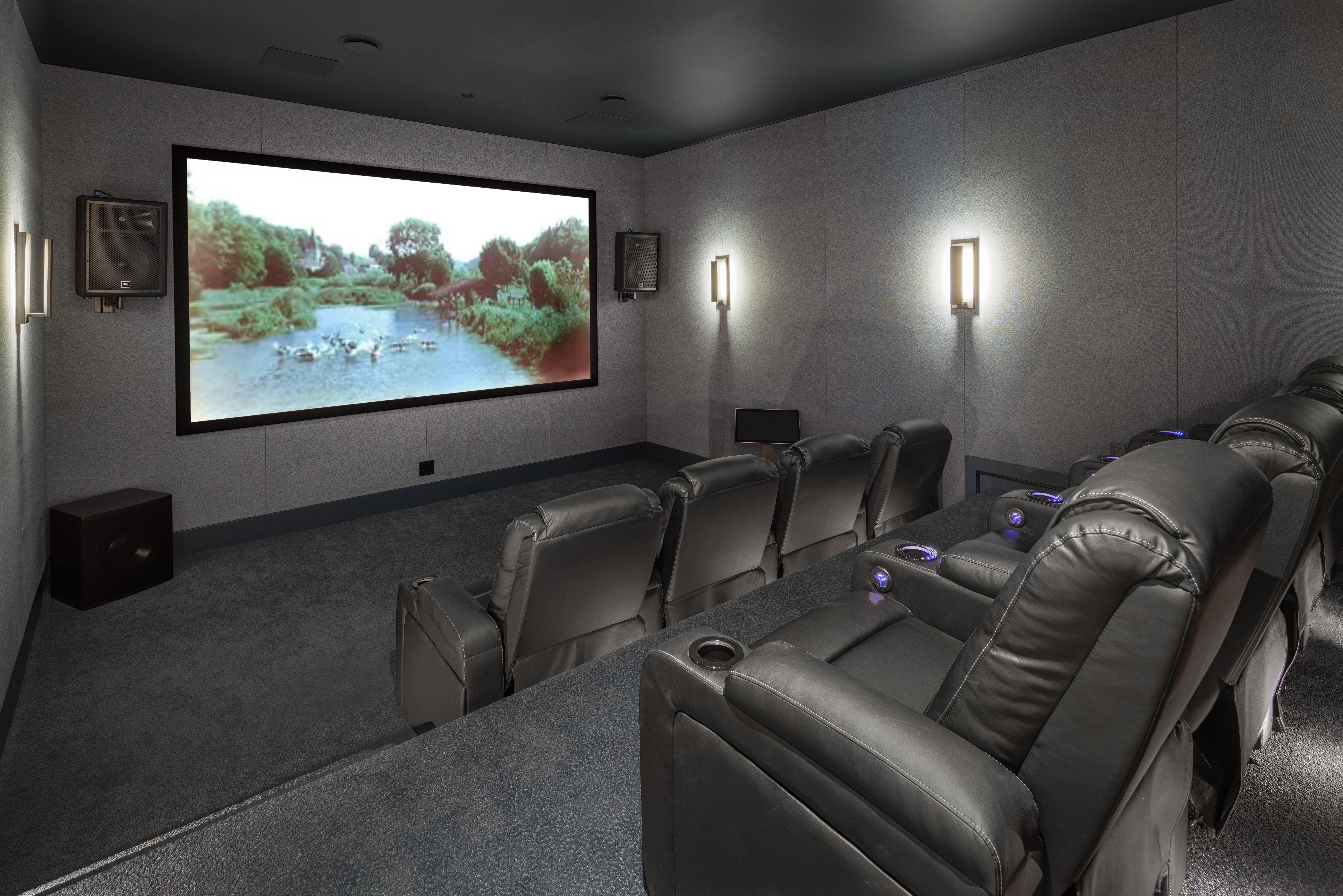 Home theater room by a BC construction company, featuring black leather recliners facing a large screen displaying a nature scene.