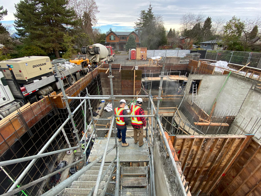 Two construction workers in safety gear stand on scaffolding at a major building site for a construction company in British Columbia, surrounded by nearby houses and trees