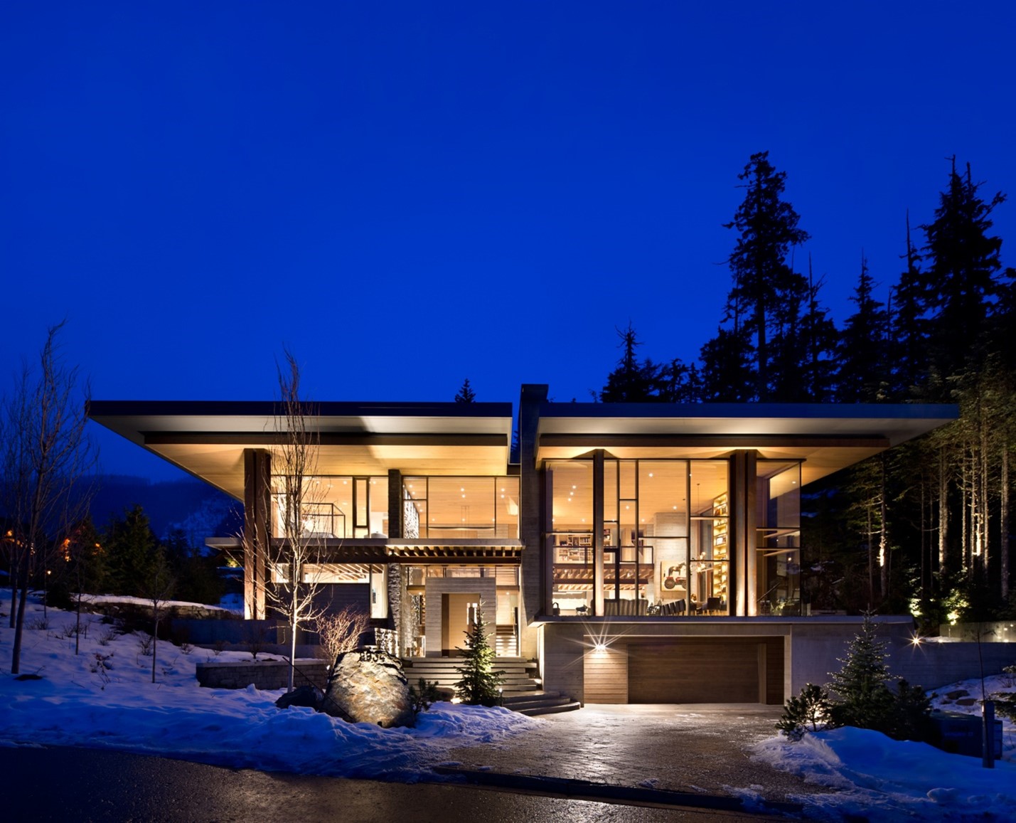 A glass-walled house lit up at night, surrounded by snow and tall trees in British Columbia, built by a leading general contractor.