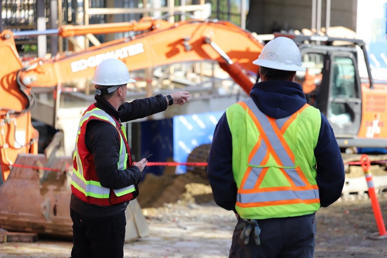 Two workers in safety gear collaborate at a job site with machinery and equipment in the background.