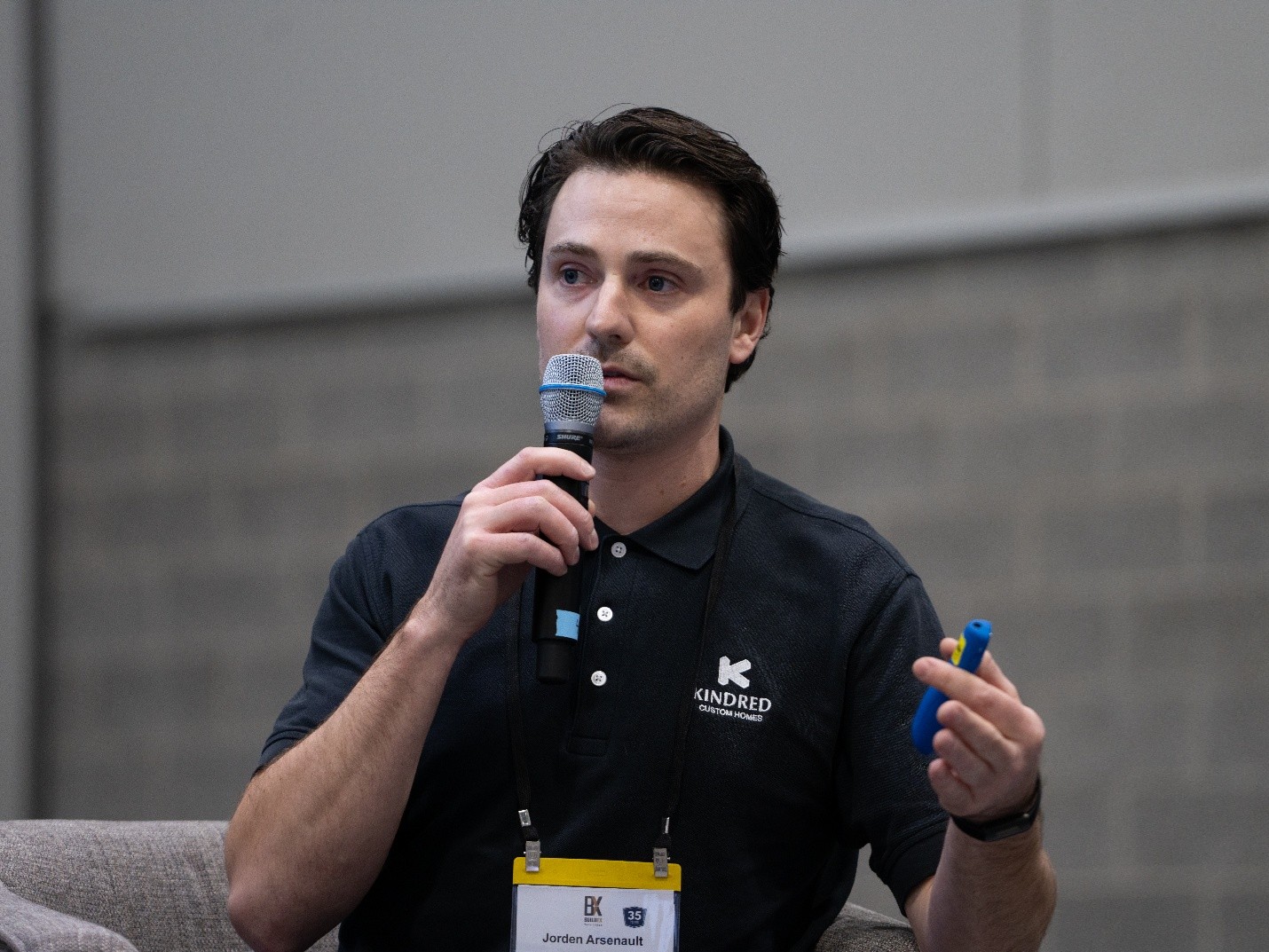 Man in a black polo shirt speaks into a microphone while holding a blue object during a panel discussion.