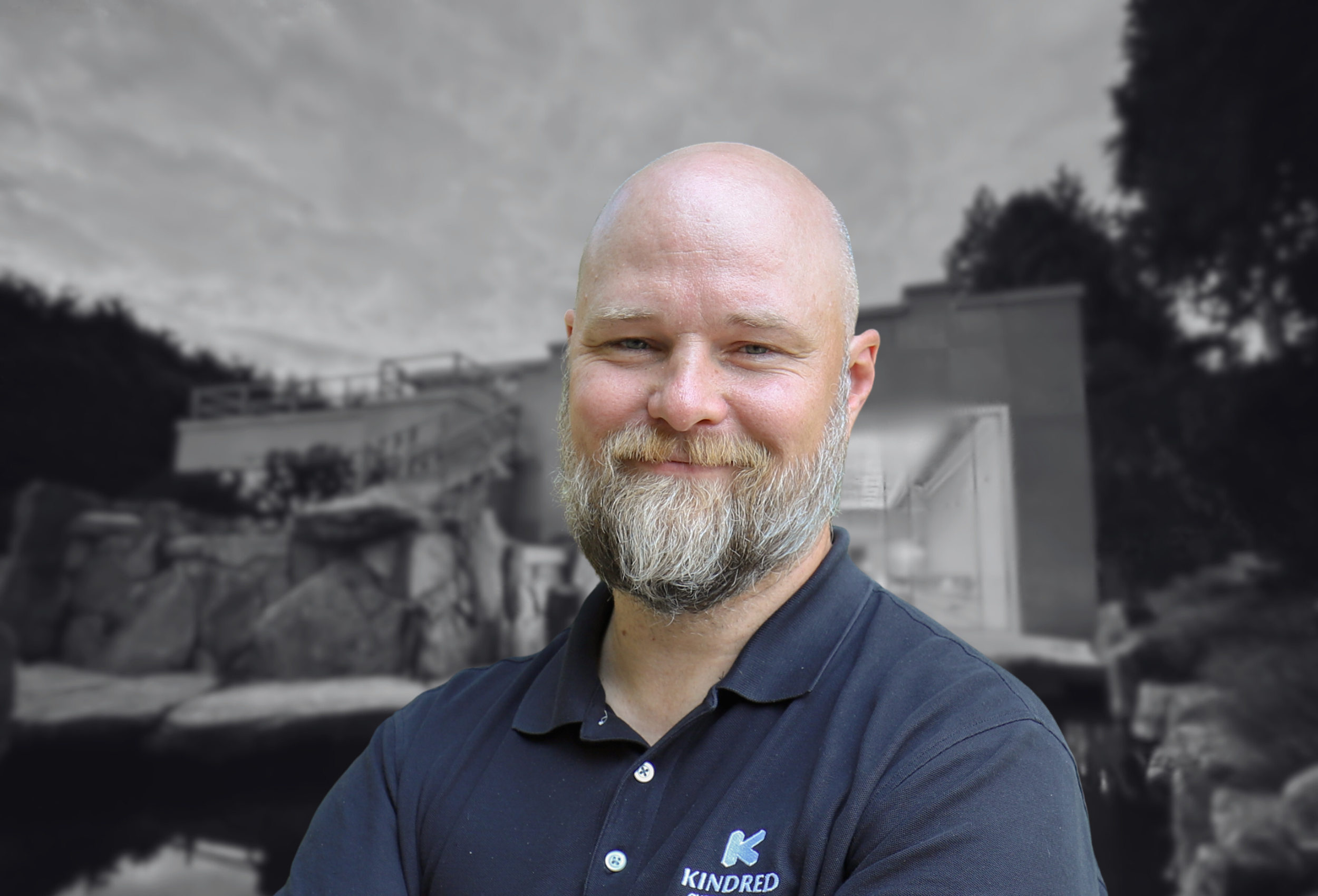 Bald man with a beard smiling, wearing a Kindred shirt, stands before a blurred outdoor building scene.