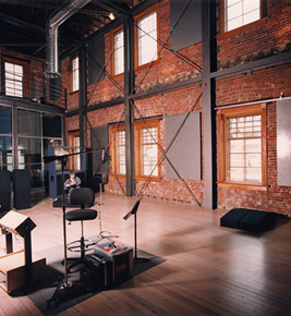 A modern recording studio with exposed brick walls, large windows, and music equipment on a wooden floor.