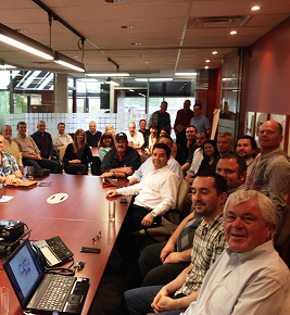 A large group of people sit and stand around a conference table in a modern office meeting room.