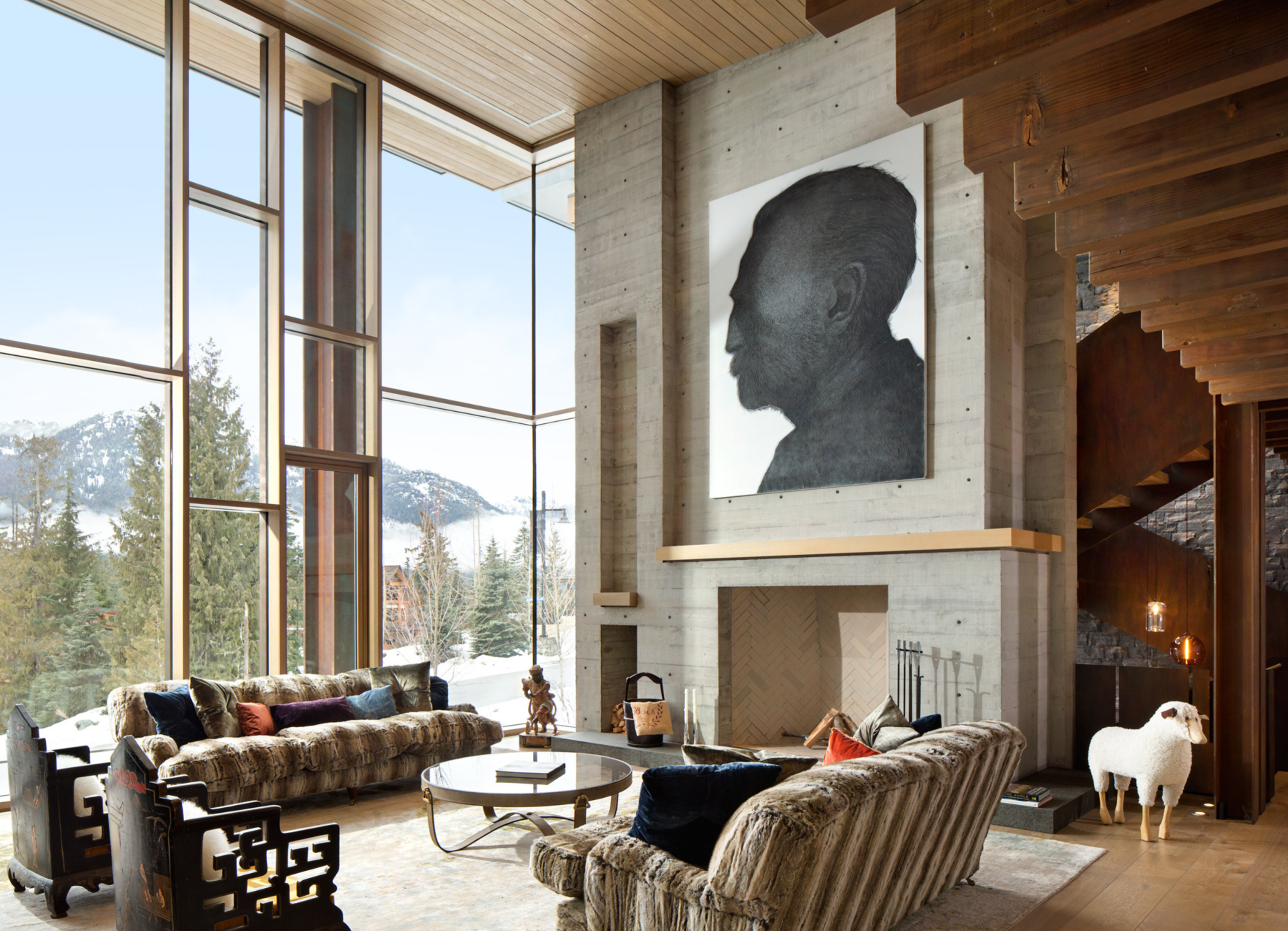 Modern living room with plush fur sofas, a large grayscale portrait on a concrete wall, and tall glass windows overlooking snowy mountains.